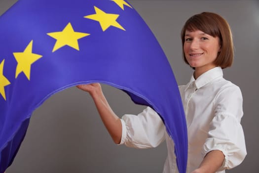 happy woman waving european flag over grey background (flash spot on at the right side above should accent the cheerful character of the image)