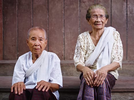 Portrait of two old asian women sitting against brown wall and looking at camera