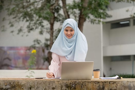 A smiling Muslim woman in a hijab studies with her laptop in a natural setting, representing her determination and growth in academics.
