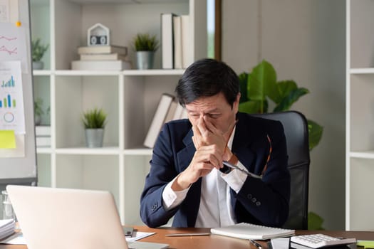 An elderly professional appears stressed while reviewing financial data on a laptop in a contemporary office space.