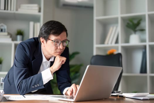 An elderly businessman ponders over data displayed on his laptop, demonstrating focus and concentration in his work.
