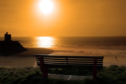 ballybunion bench in winter with view of beach castle and cliffs as the sun sets and a man jogging