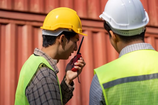 Two construction engineers in safety gear review plans while discussing logistics using a walkie-talkie.