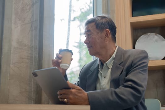 An elderly man holds a coffee cup and a tablet, looking out the window in a cafe, blending leisure with technology.