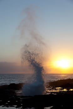 Setting sun illuminates spout of water from lava rocks on coast of Kauai