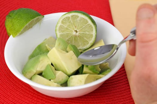 Hand about to spoon up a bite of a simple dish of just cut up avocado with lime juice from a bowl  with lime halves on and in it.