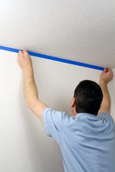 Adult male pressing blue painter&#039 s tape onto a white wall below a ceiling in order to protect the wall when he paints the ceiling above.