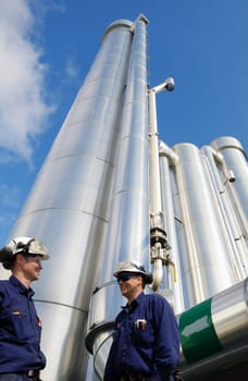 two engineers, workers with large shiney gas pipes in background
