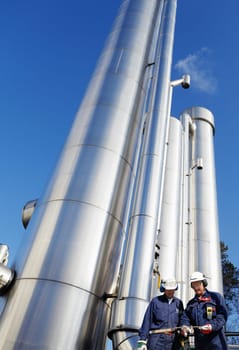 two refinery-workers standing in front of large shiney oil and gas pipes