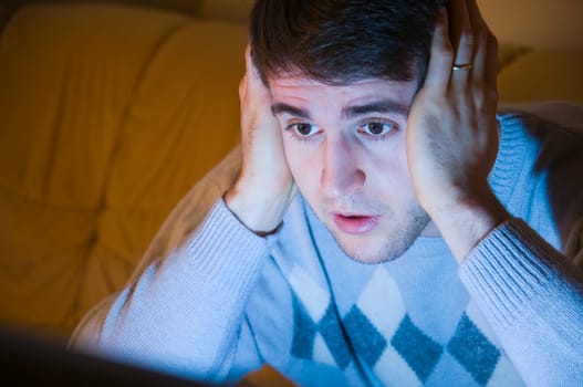 Head and shoulders shot of a young man with hands on his cheeks looking at the TV in the darkened living room