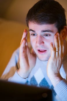 Head and shoulders shot of a young man with hands on his cheeks looking at the TV in the darkened living room