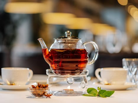 Glass teapot with warmer and candle, with hot herbal tea on table on restaurant background with bokeh. Tea concept with transparent glass teapot and cups on table in restaurant interior.