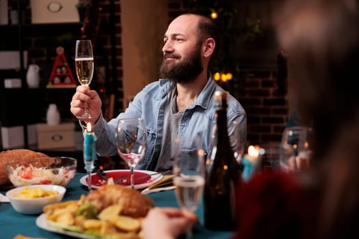 Young man raising glass for toast during christmas dinner at home, gathering with friends and family to eat traditional food and give speech at table. Joyful person enjoying festive december celebration.