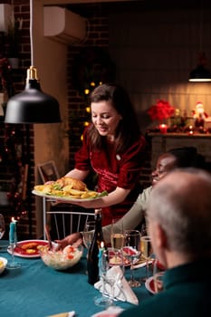Woman serving festive meal at table, celebrating christmas dinner with diverse friends and family at home. People feeling joyful eating homemade food and drinking wine during xmas eve event.