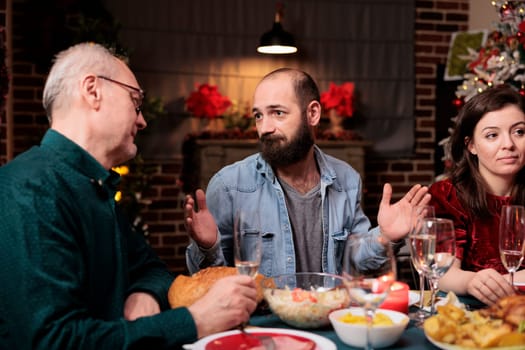 Diverse persons enjoying christmas eve holiday at home, sitting around festive dinner table and eating delicious food. Family and friends having fun sharing memories, xmas event celebration.