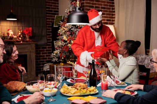 Santa claus giving gifts to persons at dinner table, spreading christmas positivity and enjoying winter celebration. Adult dressed in costume and hat offering presents to friends and family.