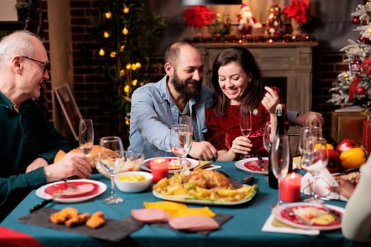 Cute couple meeting family at dinner on christmas eve holiday, gathering around table with persons to eat food and drink wine. Young adults talking to relatives during december event at home.