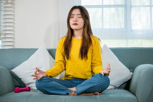 Young woman practise yoga and meditation in lotus position, mindfulness lifestyle, Asian female sits on sofa at home and chilling do yoga exercise with couch lotus pose eyes closed, healthy care