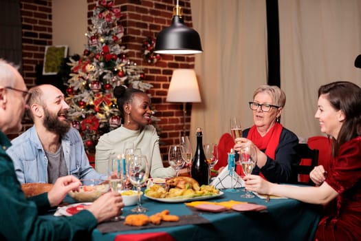 Grandmother does toast with champagne at dinner table with friends and family, enjoying christmas eve celebration at home. Senior person giving speech with raised glass for cheers, winter event.