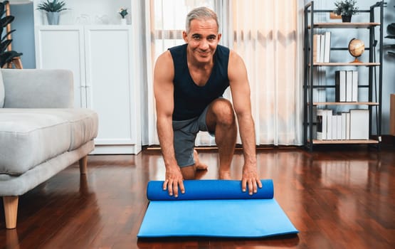 Active and sporty senior man preparing, rolling fitness exercising mat on living room floor at home. Home exercise as concept of healthy fit body lifestyle after retirement. Clout