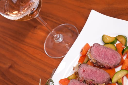 Saddle of lamb fillet with mustard and bread crust on glazed carrots and zucchini and a rosé wine in a wine glass