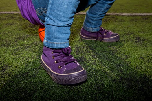 Closeup of a child sitting on a basketball with jeans and sneakers on an outdoor court