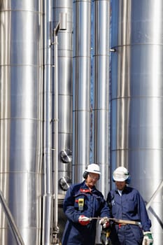 two engineers, workers with giant gas and oil pipes in background
