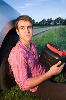 young man sits on the side of the road in the grass at the front wheel of a silver-gray car and handles a black gasoline canister