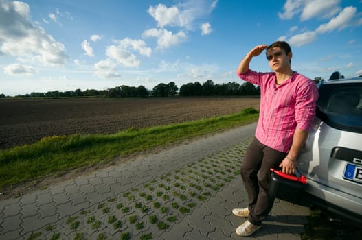 Young man with black reserve canister on a lonely country road leans helplessly against a car and waits for help