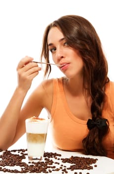 Young woman sitting at a table with a glass of latte macchiato coffee with spoon in mouth against white background