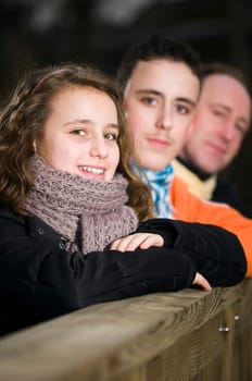 Upper body shot of a young girl in winter casual clothes on a wooden bridge railing with brother and father out of focus in the background at nightfall