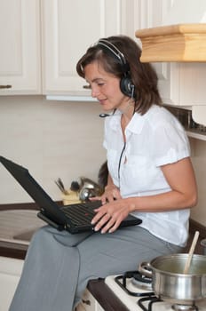 Young business person with headset working from home on laptop while cooking meal in kitchen
