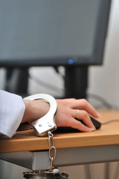 Detail of business person hand tied with handcuffs to workplace, keyboard and monitor in background.