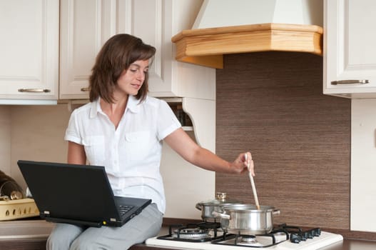 Young woman cooking meal in kitchen according to receipt from laptop (internet)