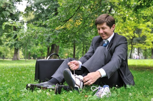 Senior business man changing working shoes for sports shoes in park (siting on grass)