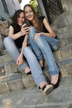 Youth lifestyle - two smiling friends (girls) siting on stairs outdoors with mobile phone