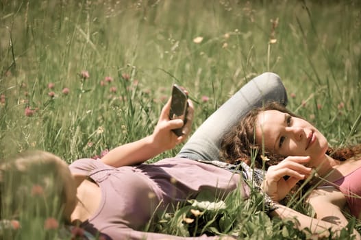 Two friends (teenage girls) relax lying on grass outside in nature - one girl is using cellphone