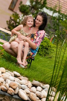 Mother with daughter siting on bench in garden - family house in background