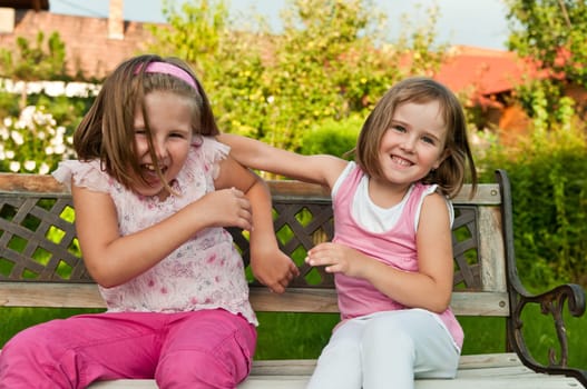 Two small girls (sisters) larking in backyard sitting on bench