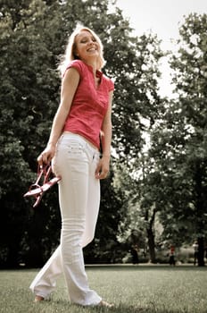 Smiling young woman holding her shoes and walking barefoot on grass in park