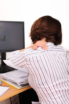 Business woman with neck pain. Focus on hand on neck with blurred monitor and documents on table.
