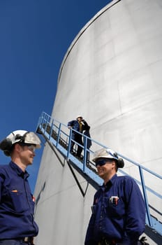 three engineers, workers, with large fuel-storage tower in background