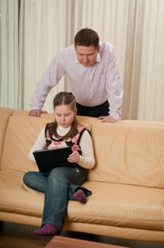 Little child (cute girl) is playing with tablet at home while her father is watching over shoulder