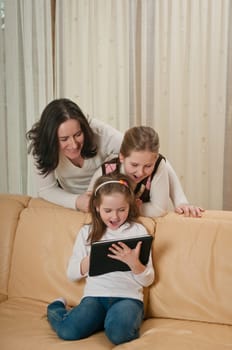 Little child is playing with tablet at home while her mother and sister are watching over shoulder