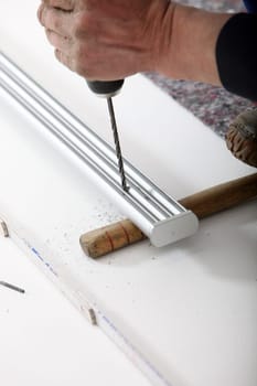 An artisan&#039 s hand using a portable cordless drill to drill a hole in a length of aluminium track supported at ground level.