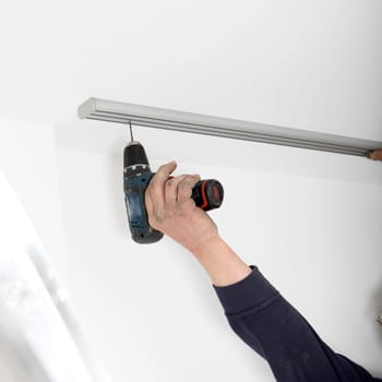 Tha arm of an artisan using a portable cordless drill to fit a length of aluminium curtain track to the ceiling
