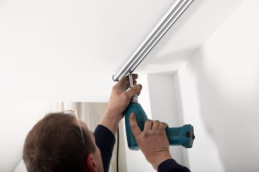 Back head view of a man using a portable drill to fit a length of metal cutain track to the ceiling