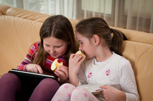 Children (sisters) playing with tablet at home on sofa and eating apple