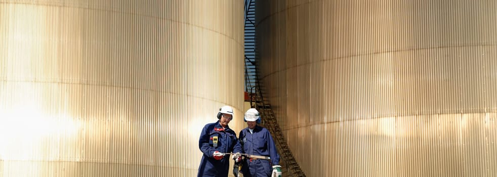 two oil and gas workers, large shiney fuel-storage towers in background, panoramic view