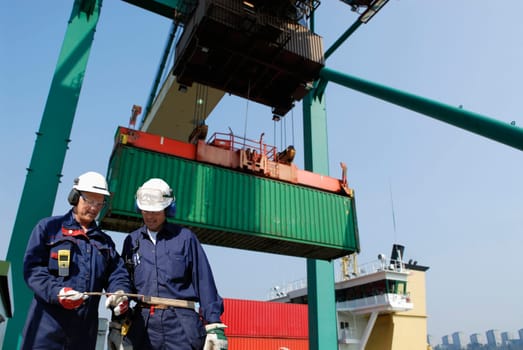 two port-workers with large crane hoisting containers in background